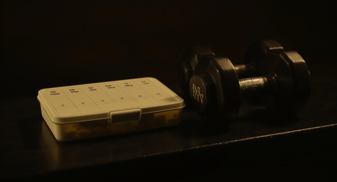 A weekly pill organiser and dumbbells side by side on a dark bench