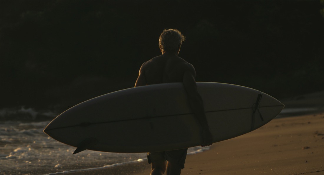 A man in his fifties carrying a surfboard toward the water at sunrise